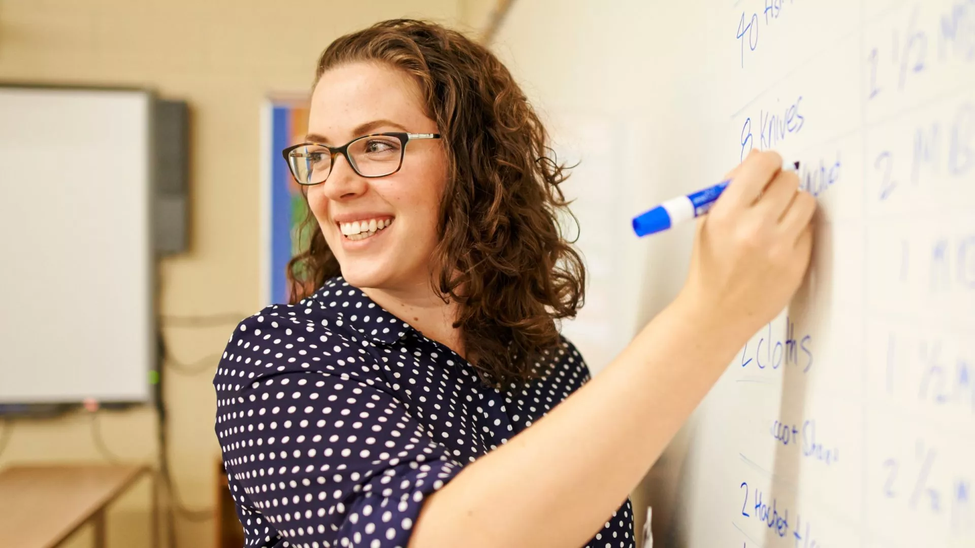 A teacher using a blue marker on a white dry erase board smiles back at her classroom