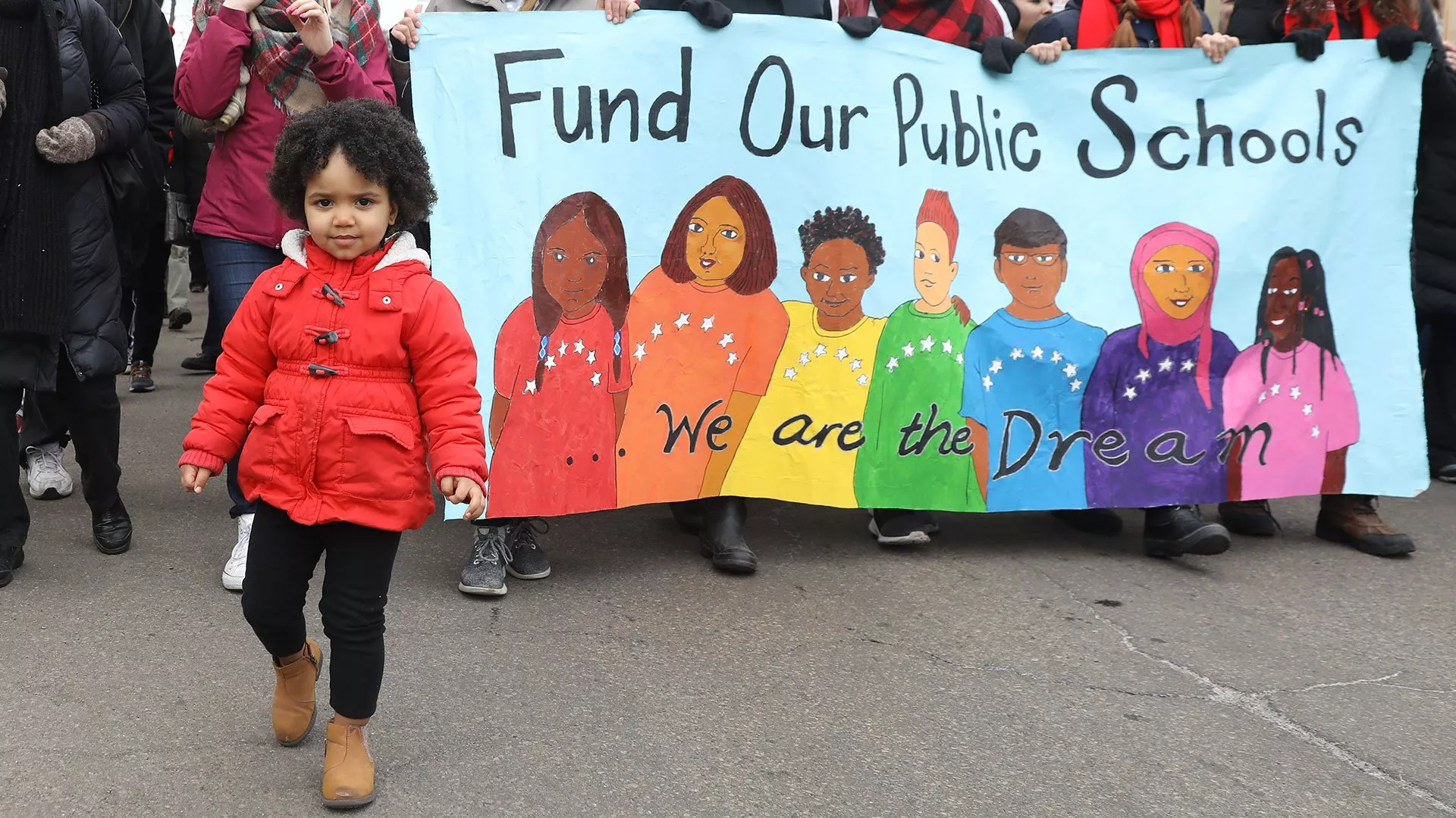 A small child standing in front of a banner that says "Fund our Public Schools." There are drawings of children on the banner with the words "We are the dream" on top of them.
