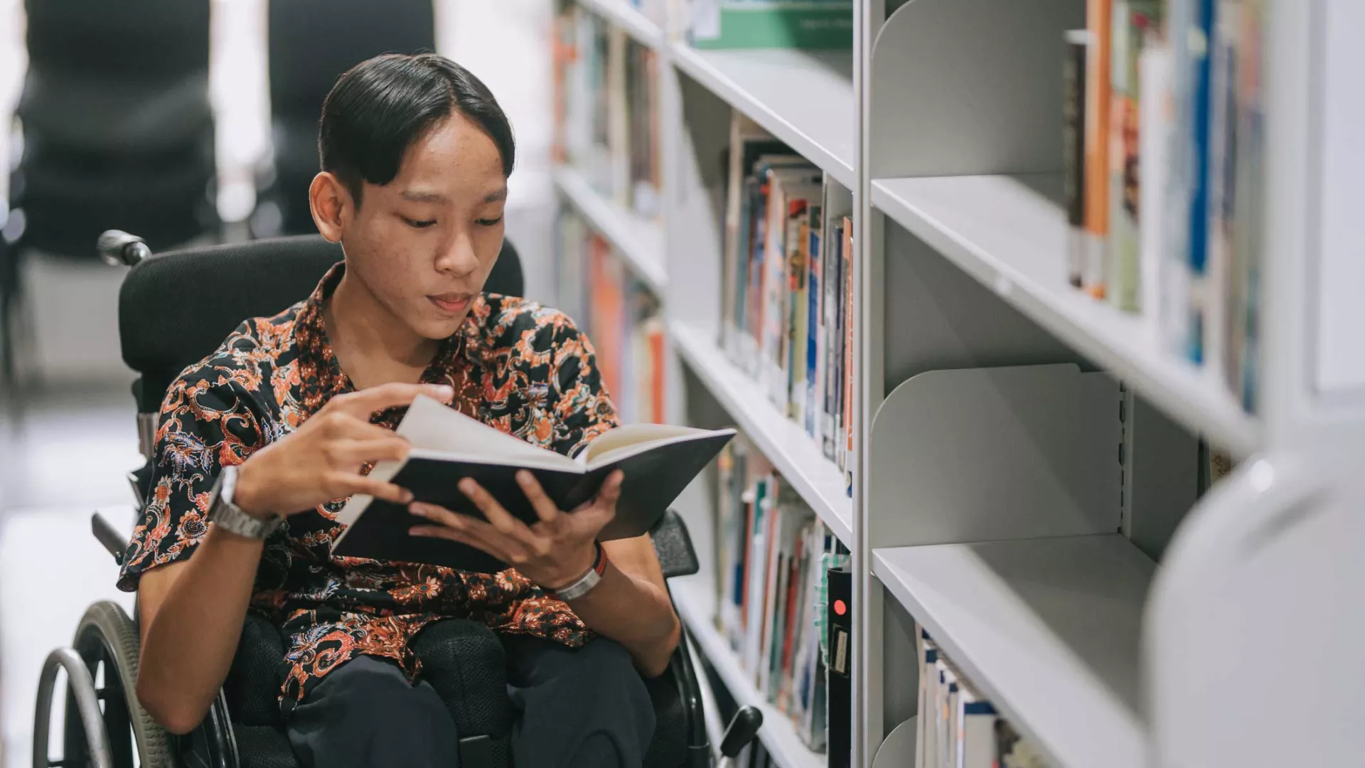 A teenage boy in a wheelchair browses books in a school library
